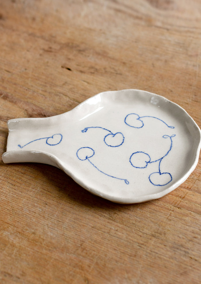 A handmade ceramic spoon rest with hand-drawn cherries in blue.