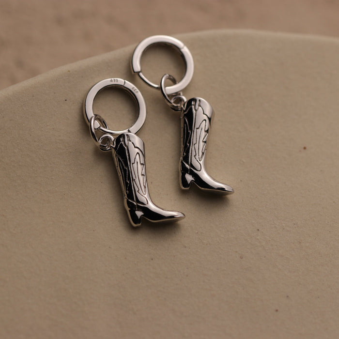 Silver cowboy boot earrings on a beige surface