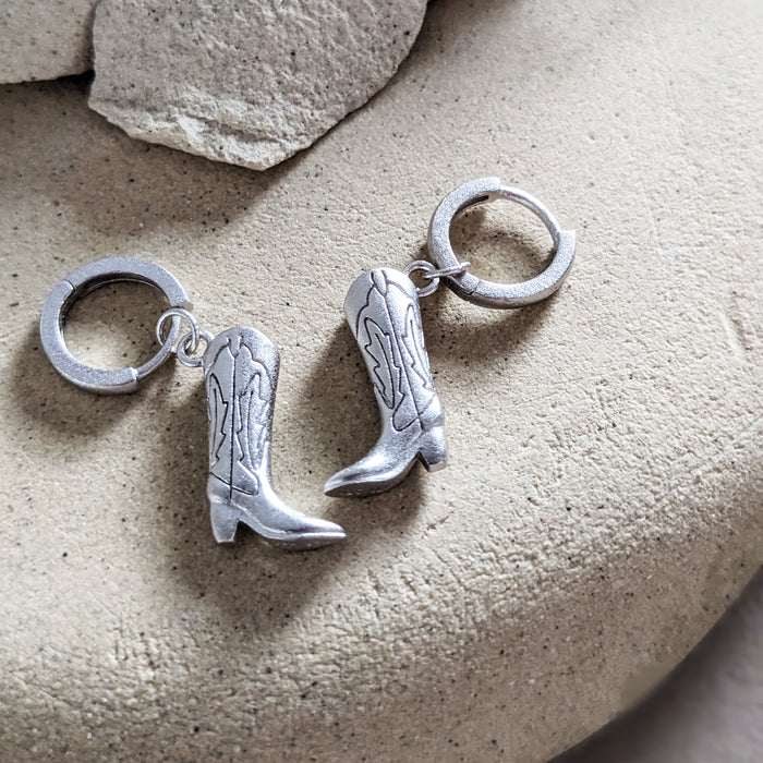 A pair of silver hoop earrings designed in the shape of cowboy boots, displayed on a beige stone surface.
