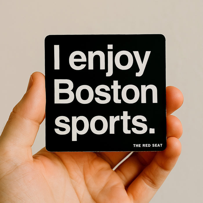 Hand holding a black square magnet with 'I enjoy Boston sports' text on a beige background