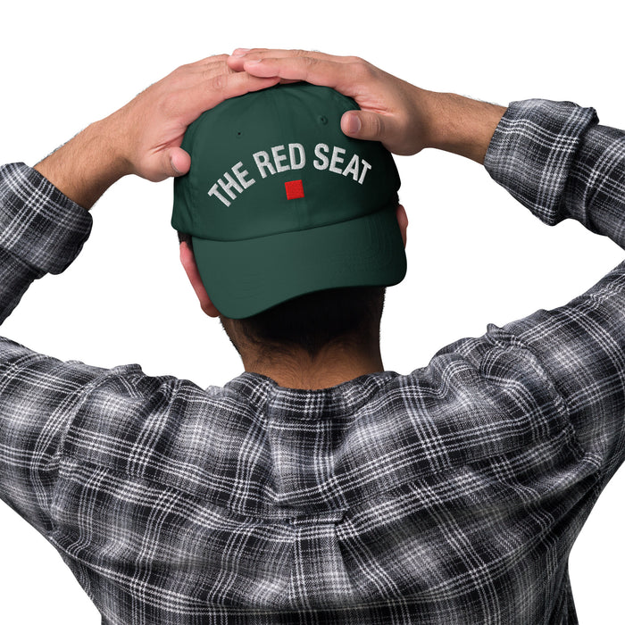Person wearing a green cap with 'The Red Seat' text, holding it on a white background